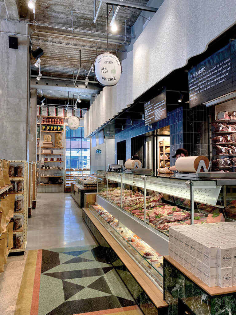 Display counters anchor the market with bakery and butchery ingredients under a white felt proscenium.
Photo credit: Stephanie Palmer